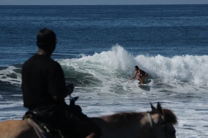 A horse rider on the beach watching a surfer catch a wave at Mizata